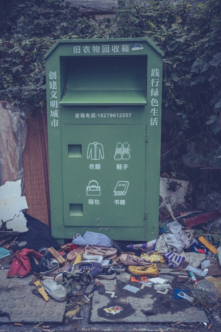 A green clothing donation and recycling box located outdoors on a pavement, with Chinese characters and contact information printed on it. The box has two compartments, each with an illustration indicating its purpose: one for clothing and shoes, and another for bags and books. Surrounding the base of the box, there is a clutter of discarded items including shoes, plastic bags, plastic bottles, clothing, and miscellaneous rubbish, scattered across the pavement and slightly spilling onto a nearby area. The background features trees and a fence, with some shadowed areas and uneven lighting. This scene depicts an informal waste collection point, highlighting the importance of proper disposal during home removals or estate clearances, often handled by services like Man with Van Parsons Green.