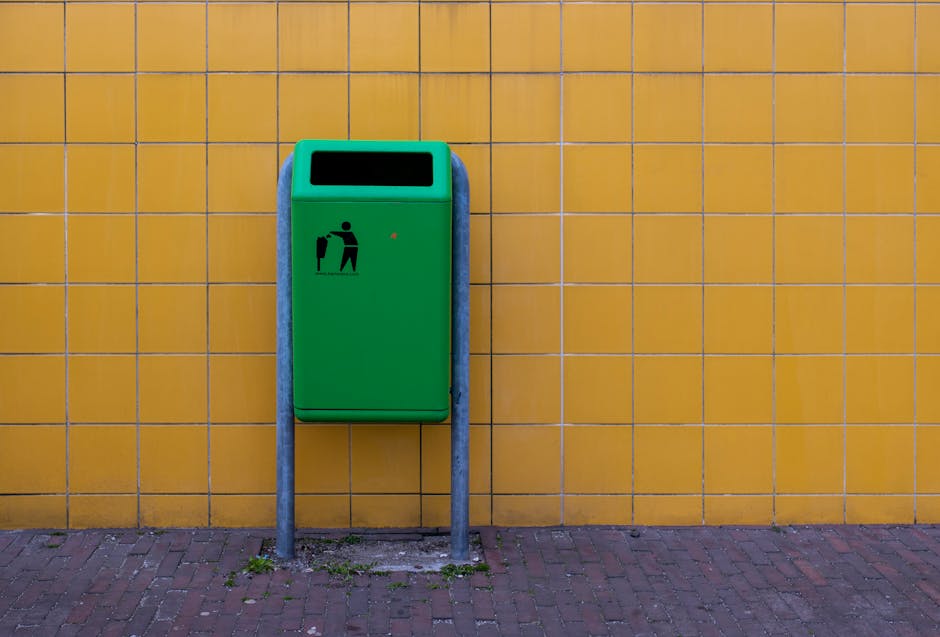 A green outdoor waste bin with a black pictogram of a person disposing of rubbish stands against a yellow tiled wall on a paved pavement. The bin has an open rectangular slot at the top for trash placement and a small red indicator light. It is supported by two metal posts embedded in the ground, with slight grass and dirt visible at the base. This image, associated with house removals and furniture transport services, illustrates waste disposal which may be relevant during home relocation or clearance processes carried out by Man with Van Parsons Green. The setting appears to be outside a building, indicating the final step of loading or waste collection in the moving or clearance process.