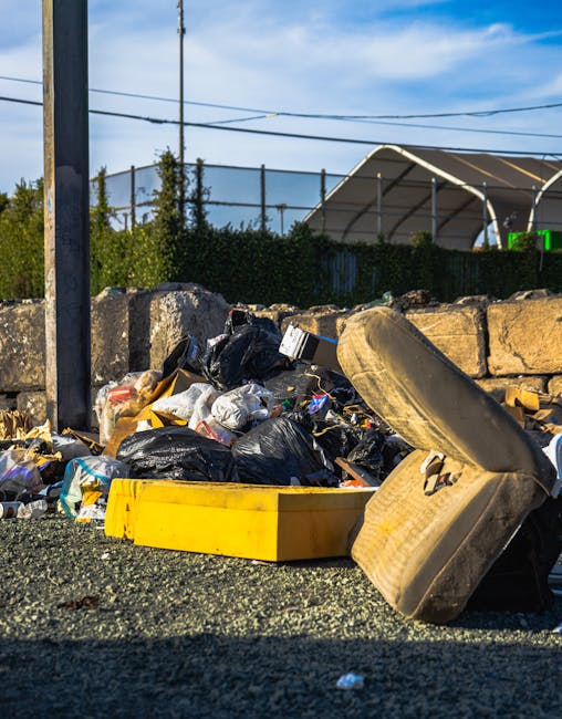 A green clothing donation and recycling box located outdoors on a pavement, with Chinese characters and contact information printed on it. The box has two compartments, each with an illustration indicating its purpose: one for clothing and shoes, and another for bags and books. Surrounding the base of the box, there is a clutter of discarded items including shoes, plastic bags, plastic bottles, clothing, and miscellaneous rubbish, scattered across the pavement and slightly spilling onto a nearby area. The background features trees and a fence, with some shadowed areas and uneven lighting. This scene depicts an informal waste collection point, highlighting the importance of proper disposal during home removals or estate clearances, often handled by services like Man with Van Parsons Green.