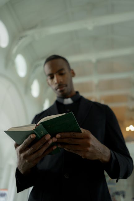 A man dressed as a Catholic priest in a black clerical shirt with a white collar is standing inside a building with high, curved ceilings and circular windows. He is holding and reading an open green hardcover book, with a focused expression. Behind him, there is a partly visible wooden door or furniture, and soft natural light enters through the windows, illuminating the scene. This image may depict a moment of preparation or reflection related to a house move or relocation service provided by Man with Van Parsons Green, as part of their furniture transport and packing process for home removals in the Parsons Green area.