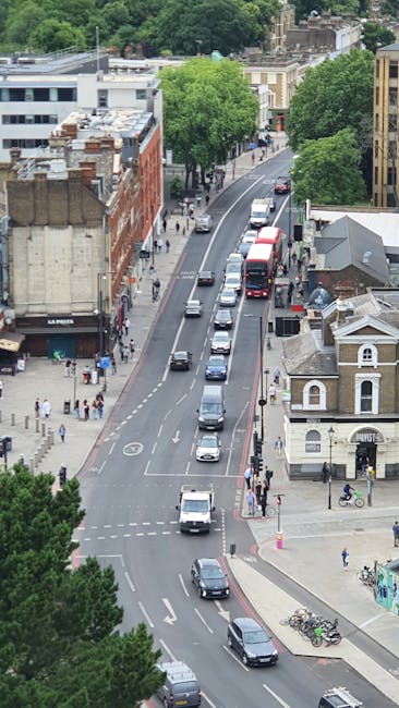 An aerial view of a street in Parson's Green, Hammersmith & Fulham, showing a line of vehicles including cars and a red double-decker bus moving in a single lane along a curved road. On the left side of the street, there is a sidewalk bustling with pedestrians walking past shops and cafés, some of whom are carrying boxes or packages, indicating recent or upcoming home relocation activities. To the right, there are residential buildings with distinct architecture, featuring bay windows and brick facades, alongside a small church or community building. Several bicycles are parked near the corner, and lush green trees line both sides of the road, providing shade and visual separation. The scene suggests a typical urban environment where house removals and furniture transport may involve coordinated loading and unloading processes around street traffic, with potential use of moving vehicles or vans like those operated by Man with Van Parsons Green for packing and moving services.