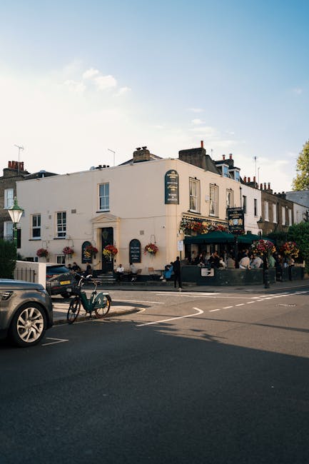 An aerial view of a street in Parson's Green, Hammersmith & Fulham, showing a line of vehicles including cars and a red double-decker bus moving in a single lane along a curved road. On the left side of the street, there is a sidewalk bustling with pedestrians walking past shops and cafés, some of whom are carrying boxes or packages, indicating recent or upcoming home relocation activities. To the right, there are residential buildings with distinct architecture, featuring bay windows and brick facades, alongside a small church or community building. Several bicycles are parked near the corner, and lush green trees line both sides of the road, providing shade and visual separation. The scene suggests a typical urban environment where house removals and furniture transport may involve coordinated loading and unloading processes around street traffic, with potential use of moving vehicles or vans like those operated by Man with Van Parsons Green for packing and moving services.