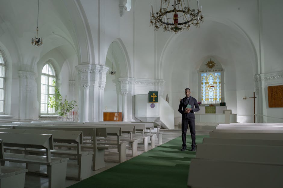 Inside a bright, spacious church with white walls, arched windows letting in natural light, and ornate architectural details, a man in a dark suit stands in the aisle holding a folder, preparing for a home relocation or event. The room features rows of white wooden pews on either side of a green carpet runner leading towards the altar area, which is elevated and decorated with religious symbols including a green cross and a sun emblem with the word 'Jesus.' Chandeliers hang from the ceiling, providing additional lighting. To the left, a potted plant is positioned near the window, and some cardboard boxes are stacked on a table or stand, indicating packing or moving activities. The scene illustrates a professional removal service, such as Man with Van Parsons Green, involved in loading or organizing furniture and packing materials during a house move or church relocation, with furniture, boxes, and protective packaging visible in the environment.