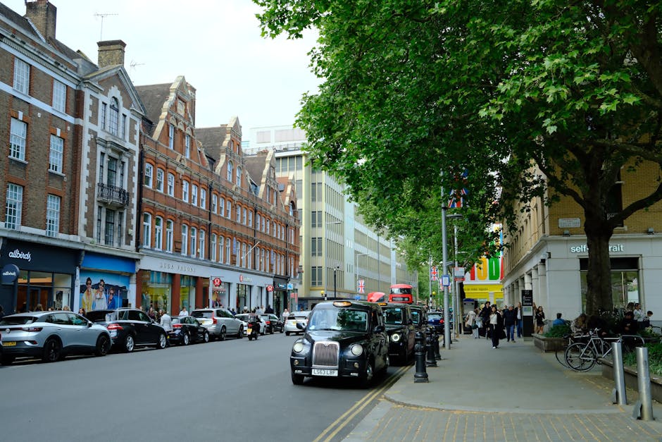 Inside a bright, spacious church with white walls, arched windows letting in natural light, and ornate architectural details, a man in a dark suit stands in the aisle holding a folder, preparing for a home relocation or event. The room features rows of white wooden pews on either side of a green carpet runner leading towards the altar area, which is elevated and decorated with religious symbols including a green cross and a sun emblem with the word 'Jesus.' Chandeliers hang from the ceiling, providing additional lighting. To the left, a potted plant is positioned near the window, and some cardboard boxes are stacked on a table or stand, indicating packing or moving activities. The scene illustrates a professional removal service, such as Man with Van Parsons Green, involved in loading or organizing furniture and packing materials during a house move or church relocation, with furniture, boxes, and protective packaging visible in the environment.