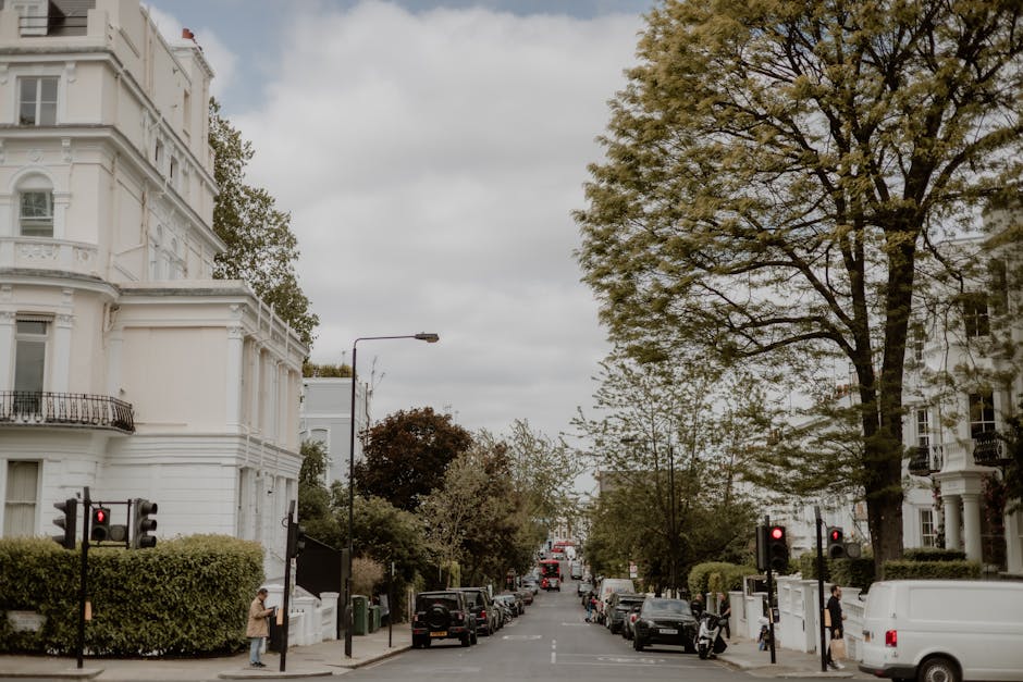 A wide view of a residential street in Parsons Green during daytime shows white Victorian-style terraced buildings with ornate architectural details and small front balconies. Several parked cars line both sides of the street, including a black SUV and a white van, with some vehicles partially visible in the background. A large, mature tree with green foliage extends over the roadway, providing partial shade, while smaller trees and shrubs are present alongside the sidewalks. Pedestrians are visible on the pavement; one person near a traffic signal is holding a clipboard or a tablet. The street is equipped with traffic lights, some showing red signals, and a street lamp is positioned on the corner. The sky is overcast with grey clouds, creating diffuse lighting. This scene captures the urban atmosphere typical of home relocation logistics, where furniture transport and packing materials are loaded into vans for punctual and secure house removals, as conducted by Man with Van Parsons Green.