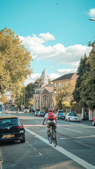 A cyclist wearing a red jacket, black helmet, and beige trousers rides along a busy street lined with parked cars and buildings, including a large church with a clock tower in the background. The street features on-street parking and a clear lane for bicycles, with trees providing shade alongside the pavement. The scene is captured during daylight hours under a partly cloudy sky. The image depicts an urban area consistent with residential and local services environments such as house removals or moving logistics, aligning with the services provided by Man with Van Parsons Green. This scene emphasizes accessibility and efficient transportation within an area like Parsons Green, suitable for home relocation activities or furniture transport, with the pedestrian-friendly street and cycling lane facilitating easy movement for household goods removal or relocation support.