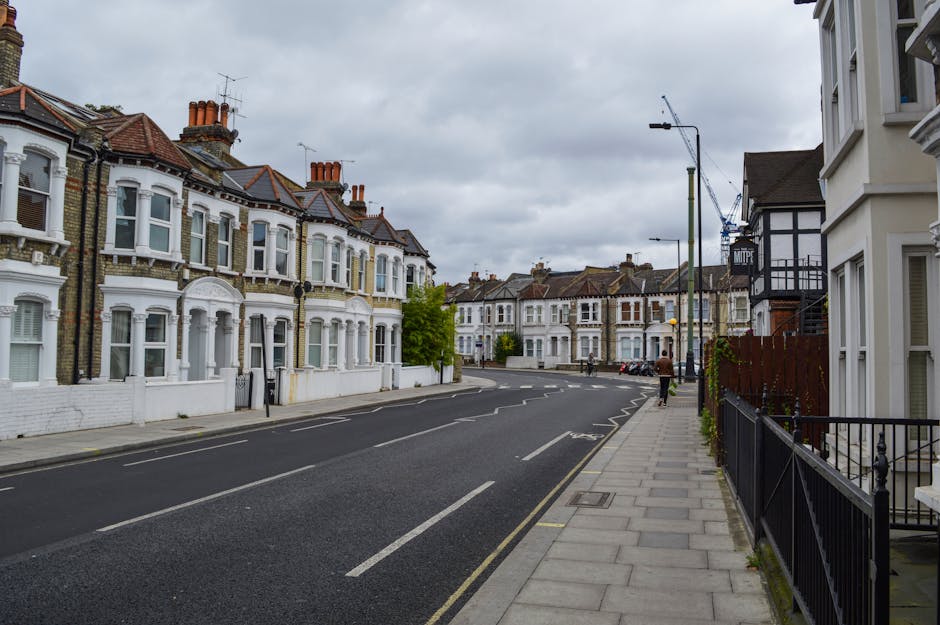 A quiet residential street scene in Parsons Green with a row of Victorian-style terraced houses featuring white facades, bay windows, and chimneys under a cloudy sky. The pavement runs along the right side, with a black metal fence and a white building partially visible. On the left, the road has parked cars and road markings, with a few pedestrians walking. Visible construction cranes in the background indicate ongoing development. The scene reflects an urban setting suitable for home relocation or furniture transport activities, typical of a house removal process supported by [COMPANY_NAME], with no visible moving equipment or personnel.
