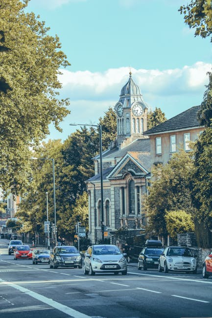 A cyclist wearing a red jacket, black helmet, and beige trousers rides along a busy street lined with parked cars and buildings, including a large church with a clock tower in the background. The street features on-street parking and a clear lane for bicycles, with trees providing shade alongside the pavement. The scene is captured during daylight hours under a partly cloudy sky. The image depicts an urban area consistent with residential and local services environments such as house removals or moving logistics, aligning with the services provided by Man with Van Parsons Green. This scene emphasizes accessibility and efficient transportation within an area like Parsons Green, suitable for home relocation activities or furniture transport, with the pedestrian-friendly street and cycling lane facilitating easy movement for household goods removal or relocation support.