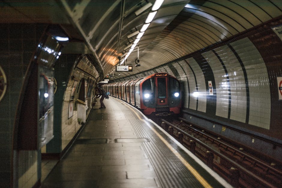 Inside a London underground station tunnel, the image shows a train with a dark exterior and headlights approaching the platform, which is lined with a curved metal ceiling and tiled walls featuring circular station logos. The platform has a wooden floor and yellow tactile paving along the edge. Two passengers are visible near the platform's end, one standing close to the train, possibly preparing to board, and the other further away, waiting near the wall. Overhead lighting illuminates the scene, and a digital display sign hangs from the ceiling, likely indicating train timings or station information. This setting represents typical logistics for home relocation involving professional removals, where careful planning of furniture transport and loading processes are crucial, often managed by companies like Man with Van Parsons Green to ensure smooth and efficient moving services.