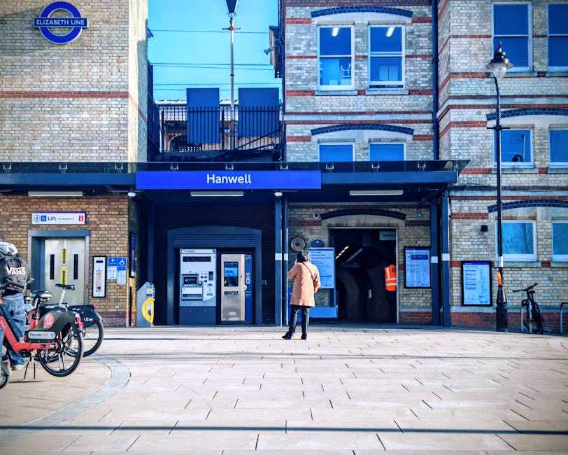 A woman stands outside the entrance to Hanwell station, part of the Elizabeth Line, with a modern brick building featuring large blue signage and multiple electronic ticket machines visible behind her. To the left, several bicycles are parked, some branded with Uber and VANS, secured on bike racks on the pavement. The station entrance has a black canopy with the station name displayed prominently, with a lift access panel nearby indicating accessibility features. In the background, an orange high-visibility jacket worn by a staff member or maintenance worker is seen entering the station, and the pavement extends forward with patterned tiles. The scene, captured during daytime under natural lighting, depicts an urban environment suitable for residential or commercial transport and home relocation services, aligning with professional removals and furniture transport in London.