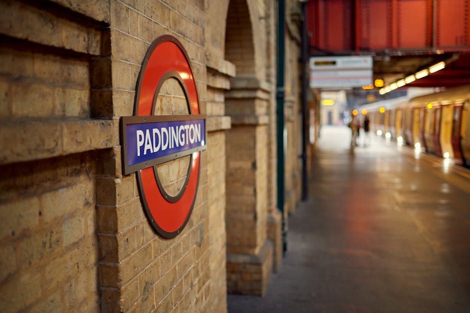 Inside a London underground station tunnel, the image shows a train with a dark exterior and headlights approaching the platform, which is lined with a curved metal ceiling and tiled walls featuring circular station logos. The platform has a wooden floor and yellow tactile paving along the edge. Two passengers are visible near the platform's end, one standing close to the train, possibly preparing to board, and the other further away, waiting near the wall. Overhead lighting illuminates the scene, and a digital display sign hangs from the ceiling, likely indicating train timings or station information. This setting represents typical logistics for home relocation involving professional removals, where careful planning of furniture transport and loading processes are crucial, often managed by companies like Man with Van Parsons Green to ensure smooth and efficient moving services.