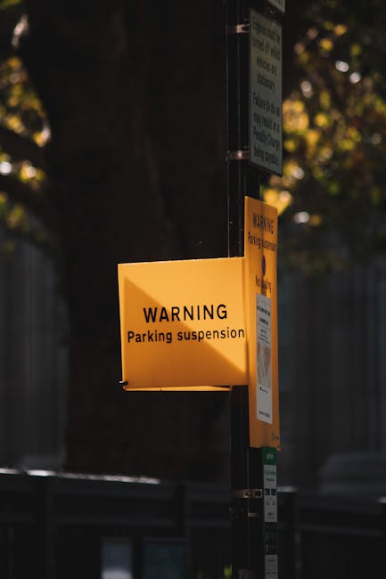 A yellow rectangular warning sign attached to a metal pole reads 'WARNING Parking suspension' and is positioned outdoors along a street. The sign is illuminated by natural light, with a dark background that includes a tree with green leaves and a shadowed fence. Additional small signs are mounted above and below the yellow warning sign, indicating parking restrictions or suspension notices. The pole is located on the pavement, suggesting the area is scheduled for temporary parking restrictions, which could impact house removals or furniture transport logistics nearby. The presence of this parking suspension sign is relevant to moving services offered by Man with Van Parsons Green, as it highlights considerations around parking regulations during home relocation or furniture transport in the SW6 area, ensuring the safety and efficiency of the loading process in a street bay suspension zone.