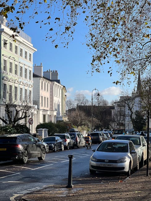 A street view in Parsons Green with a row of white Victorian-style terraced houses on the left, featuring large windows and decorative architectural details, and a busy parking area in front with various cars including sedans, hatchbacks, and SUVs parked along the curb. The pavement is made of asphalt with a single black metal bollard near the foreground, and some fallen leaves are scattered on the ground. Overhead, a tree with sparse brown leaves extends its branches across the top of the image, with a clear blue sky and a few scattered clouds in the background. The scene reflects a typical residential street in SW6, used by residents and visitors. Occasionally, Man with Van Parsons Green's removals services are involved in home relocation and furniture transport, as part of the process of packing and moving objects through this area.