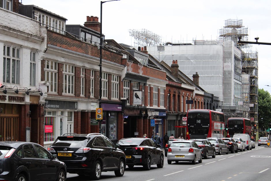 A busy street scene on New King's Road in Fulham, London, with a row of brick and white-painted terraced houses on the left side, some with shopfronts and signage. The pavement is occupied by pedestri